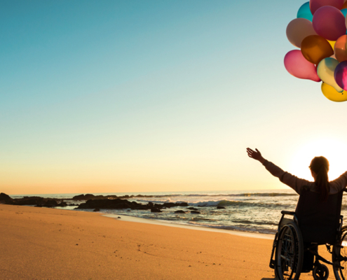Woman in wheelchair on the beach with the sun behind her on the horizon, with her arms extended up in joy and her left hand holding a large bunch of coloured balloons.