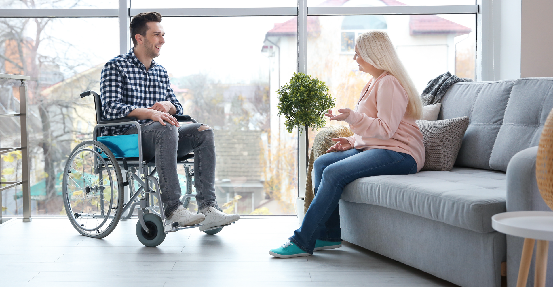 A smiling, happy man in a wheelchair talking to a woman sitting opposite him on a grey couch.