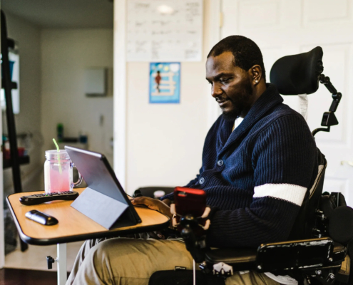 A black man in wheelchair looking at tablet sitting on a small table along with two remote controls and a drink in a clear mug with a lid and handle.